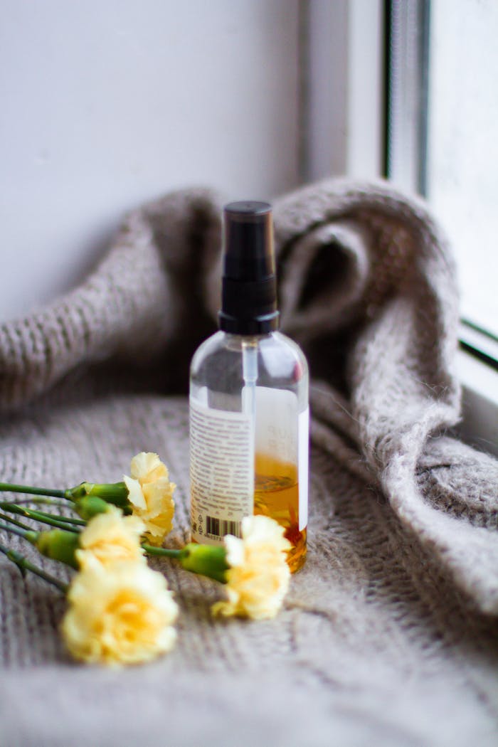 A close-up of a spray bottle beside yellow flowers on a knitted blanket by a window.